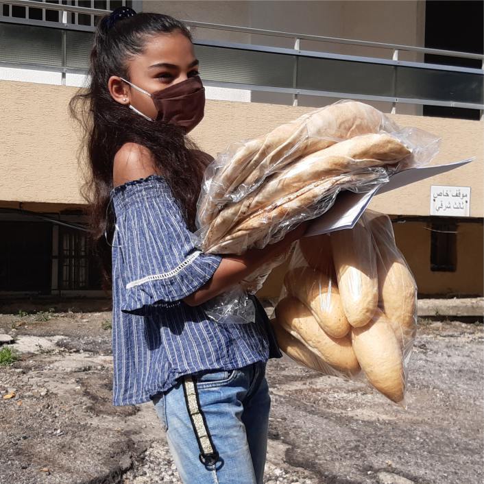 Young girl dressed in blue carrying an armful of loaves of bread.