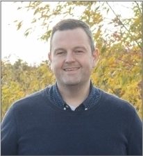 White man with short brown hair in a blue sweater smiling at camera; outdoors